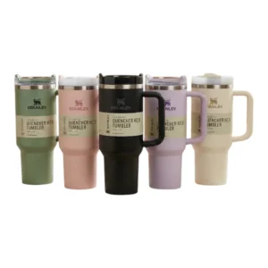 خمسة أكواب ستانلي Quencher H2.0 بألوان متعددة (أخضر، وردي، أسود، لافندر، كريمي) موضوعة في غرفة معيشة دافئة.